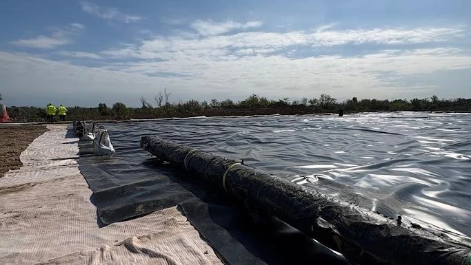 Ampia superficie coperta da un telo impermeabile nero, con due persone in giubbotto ad alta visibilità sullo sfondo