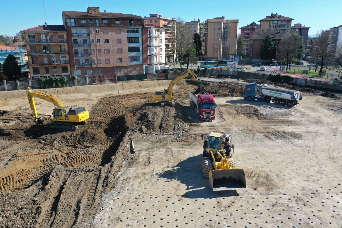 Veduta dall’alto del cantiere nell’area Ex ABB a Lodi, con escavatori, una pala gommata e autocarri tra i palazzi del quartiere