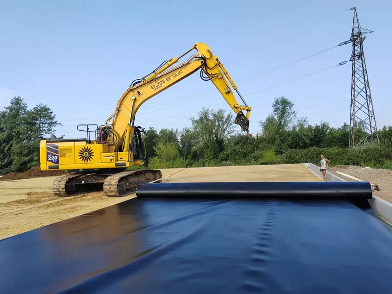 Escavatore ELIOS che stende un grande telo impermeabile nero su un piano di sabbia, con un traliccio elettrico e il bosco sullo sfondo