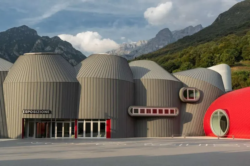 Edificio del Museo Moto Guzzi a Mandello del Lario, con volumi cilindrici in lamiera grigia e una cupola rossa davanti alle montagne