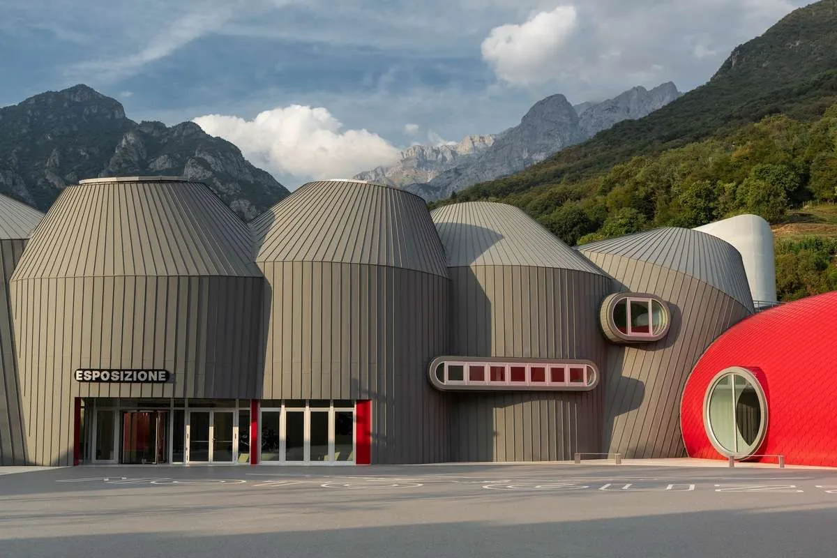 Edificio del Museo Moto Guzzi a Mandello del Lario, con volumi cilindrici in lamiera grigia e una cupola rossa davanti alle montagne