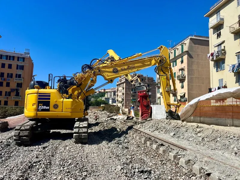 Escavatore ELIOS Ambiente con cesoia rossa sulla massicciata dei binari, tra i palazzi di Genova, con alcuni operatori sullo sfondo